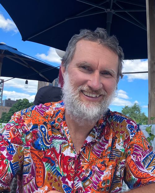 A photo of author Brett Hodnett. He is seated, his head and upper torso in view, his lower body a mystery below. With a grey beard and short messy hair, he’s smiling, his collared shirt an explosion of colours. It's sunny and the sky is blue with fluffy white clouds, though they are largely blocked from view by two patio umbrellas. Through the railing to Brett’s side is that water below? It's hard to be sure.
