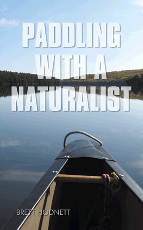 The book cover of Paddling with a Naturalist. The cover is a photograph taken from the front seat of a canoe. The nose of the canoe is in the image, the deck and carrying handle making up the foreground, ropes tied messily to them.  Extremely calm flat water is in all directions beyond, and barely perceptible sparse clouds can be seen reflecting from it. Far in the background, thickly treed gentle hills run horizontally, the dark green sandwiched between the calm blue water below, and the slightly lighter blue sky above. In bold white translucent type 'Paddling With a Naturalist' is printed over the top half of the book cover. In much smaller type below, the author, Brett Hodnett, has his name printed in the same white, translucent type.